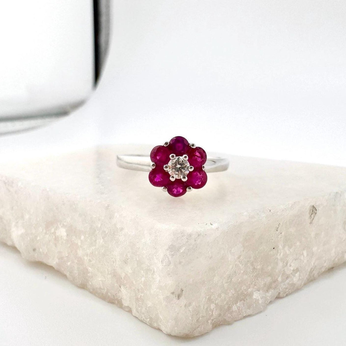 May include: A flower-shaped ring with a silver band. The ring features a cluster of vibrant red gemstones forming the petals, surrounding a central, smaller, clear gemstone. The ring is set on a white, textured surface.