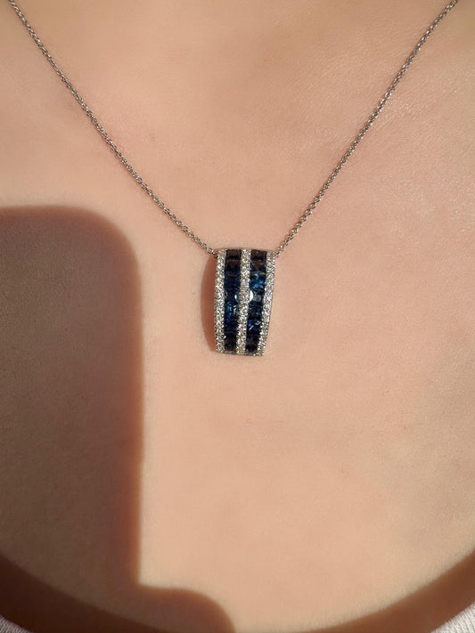 May include: A silver necklace featuring a pendant with two rows of dark blue gemstones and a row of smaller, clear gemstones. The pendant is rectangular and hangs from a delicate chain.