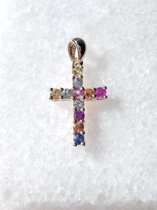May include: Multicolored sapphire cross pendant.  The cross features small, round sapphires in various colors including pink, purple, blue, and yellow, set in a silver or white gold setting. The pendant has a simple bail for hanging from a necklace chain. The gemstones are vibrant and appear to be high quality. This is a religious jewelry item.