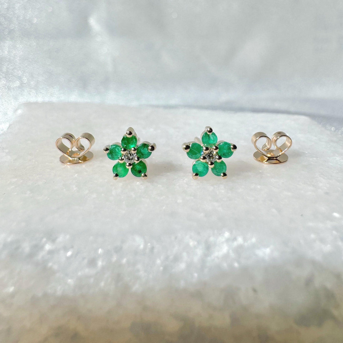 May include: A pair of gold flower-shaped stud earrings with green gemstones and a small diamond in the center of each flower. Two gold earring backs are also visible. The earrings are displayed on a white surface.