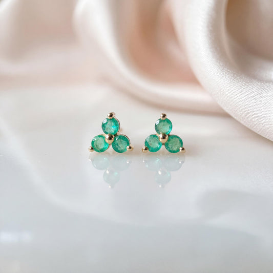 May include: A pair of stud earrings featuring three round, vibrant green gemstones arranged in a triangular cluster, set in a polished gold-tone metal frame. Each earring showcases a symmetrical design with a central stone and two side stones.