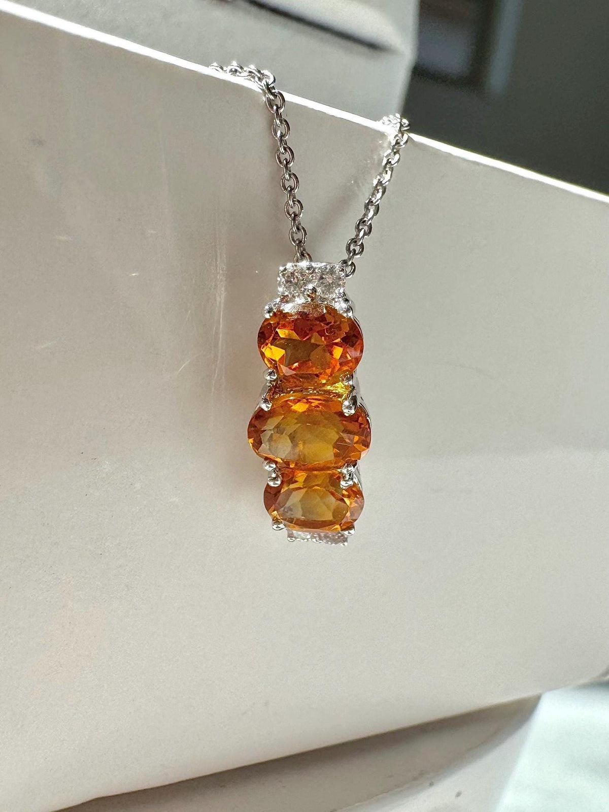 May include: A silver necklace with a pendant featuring three oval-cut, orange-yellow gemstones stacked vertically. The pendant is accented with small, clear stones at the top and bottom. The chain is delicate and silver.