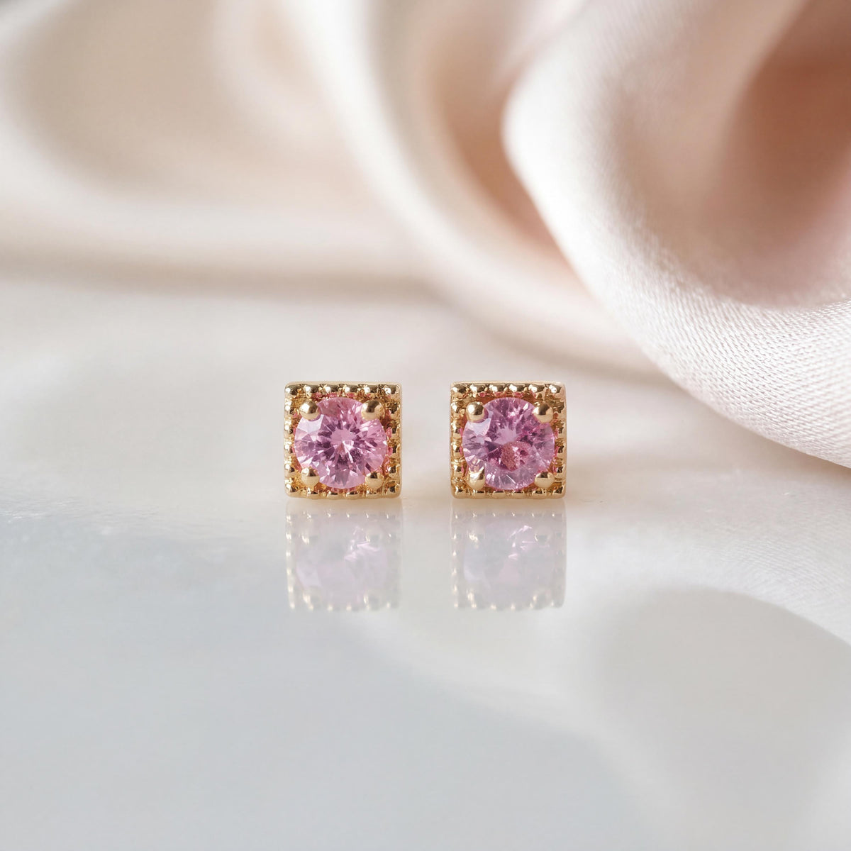 May include: A pair of stud earrings featuring round, faceted pink stones set in square, gold-toned metal frames with a beaded edge detail. The earrings are positioned side-by-side, displaying a clear reflection on the surface below them.