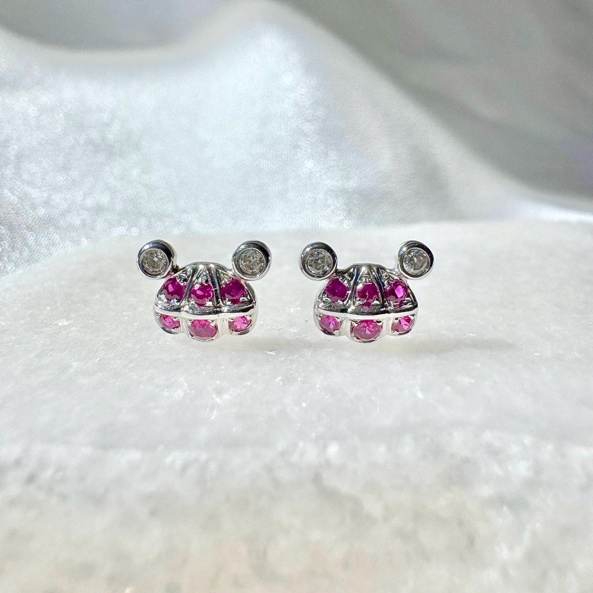 May include: A pair of silver stud earrings. Each earring features a stylized design with a central cluster of pink gemstones and two small, round, clear stones. The earrings are set against a white background.