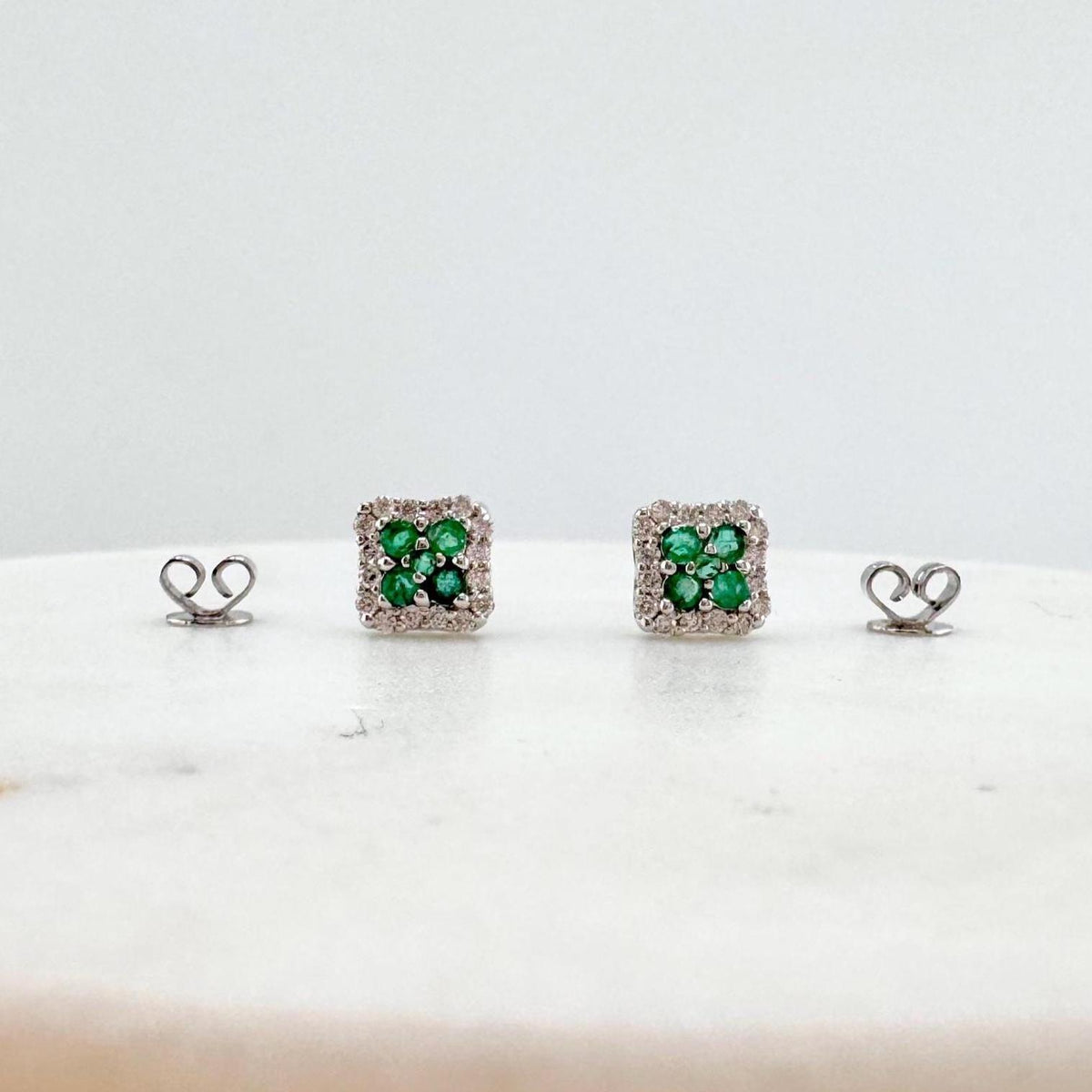 May include: A pair of square-shaped stud earrings. Each earring features a cluster of small, vibrant green gemstones surrounded by a halo of smaller, clear stones. The earrings are set on a light-colored surface.