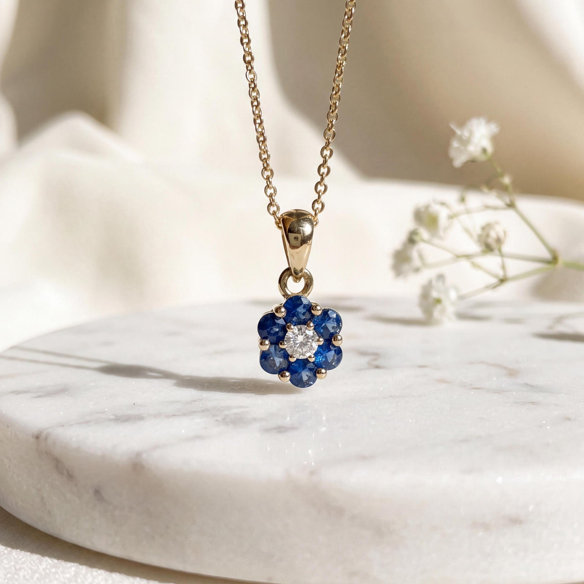 May include: A gold chain necklace featuring a flower-shaped pendant. The pendant consists of six round blue sapphire stones surrounding a single central clear white diamond, all set in a polished gold metal frame with a delicate gold link chain.