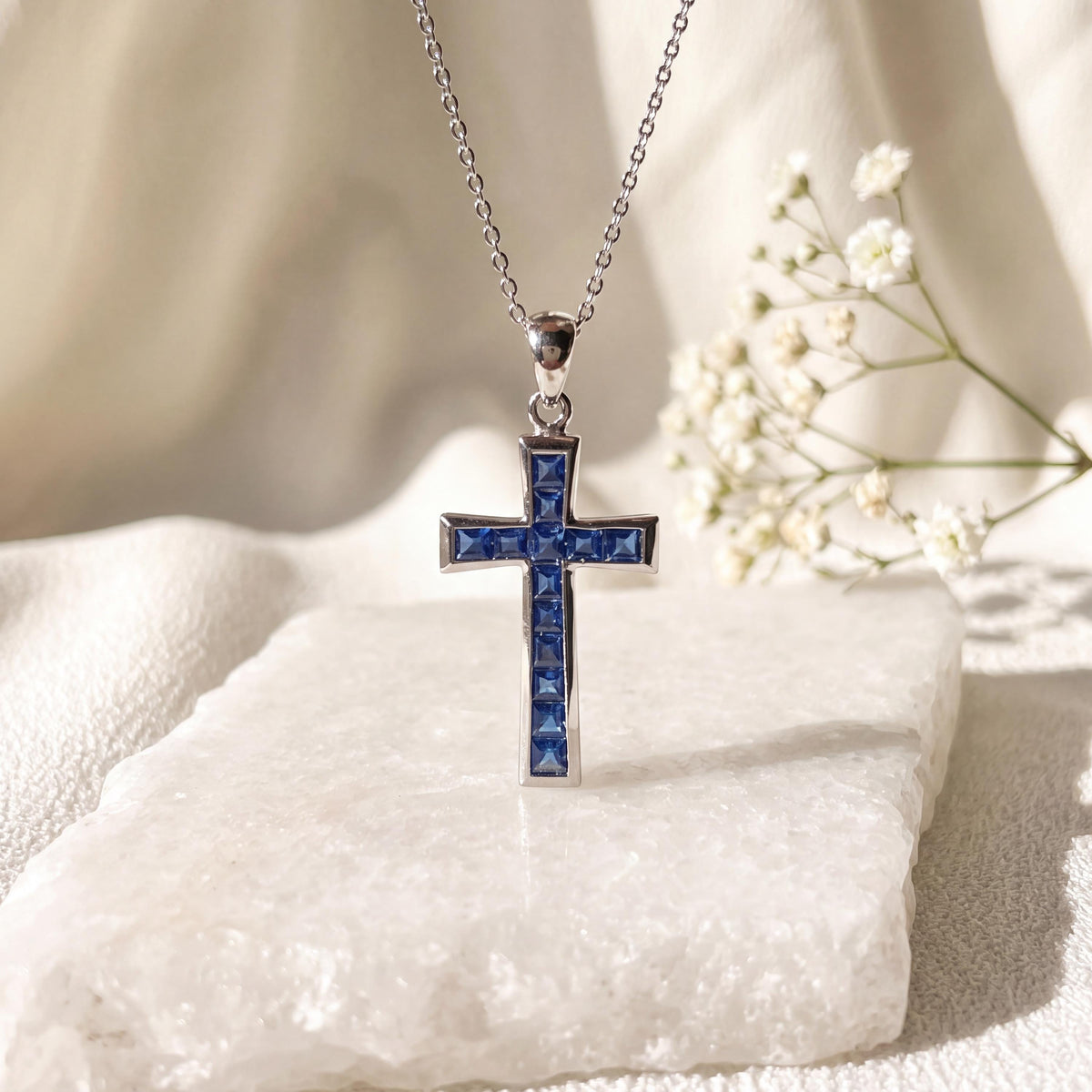 May include: A silver-toned metal cross pendant suspended from a delicate matching chain. The cross features a channel setting filled with multiple small, square-cut blue gemstones that run vertically and horizontally along the center of the piece.