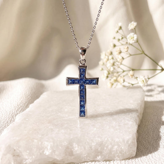May include: A silver-toned metal cross pendant suspended from a delicate matching chain. The cross features a channel setting filled with multiple small, square-cut blue gemstones that run vertically and horizontally along the center of the piece.