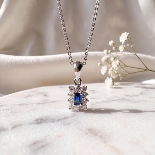 May include: A silver-toned necklace featuring a rectangular pendant with a central baguette-cut blue gemstone surrounded by a halo of small, clear, round-cut stones. The pendant hangs from a delicate silver-toned chain with a polished finish.