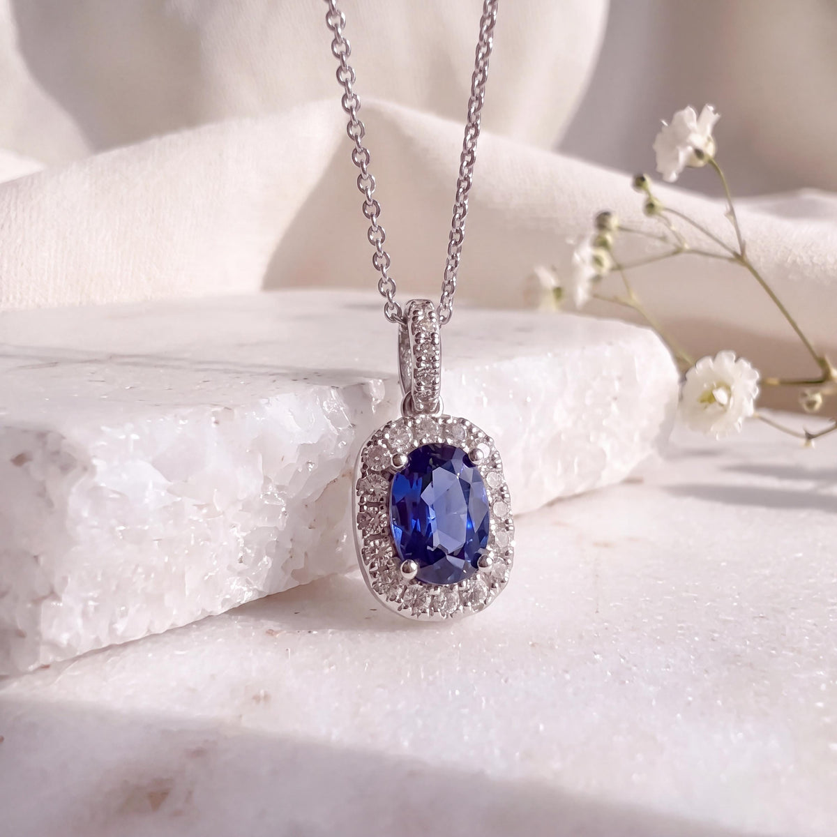 May include: A silver-toned necklace featuring an oval-shaped deep blue gemstone pendant surrounded by a halo of small clear stones. The pendant hangs from a delicate chain, with the setting and chain appearing to be made of a polished metallic material.