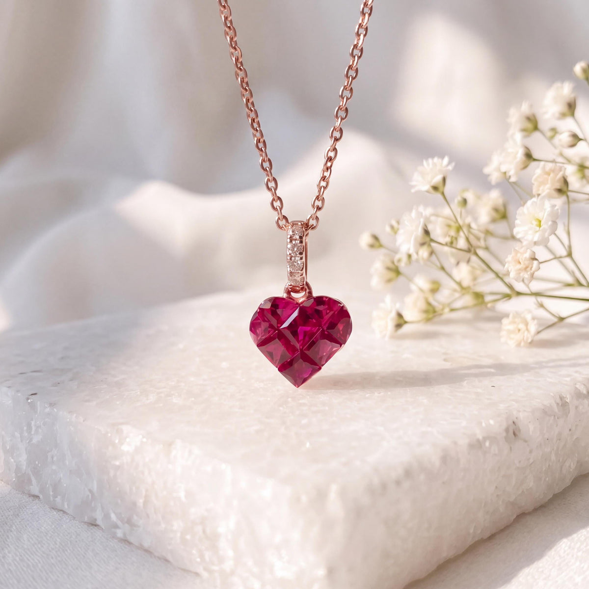 May include: A rose gold necklace featuring a faceted, deep red heart-shaped pendant. The pendant is suspended from a delicate chain and includes a small, rectangular bail adorned with tiny, clear, sparkling stones at the top connection.
