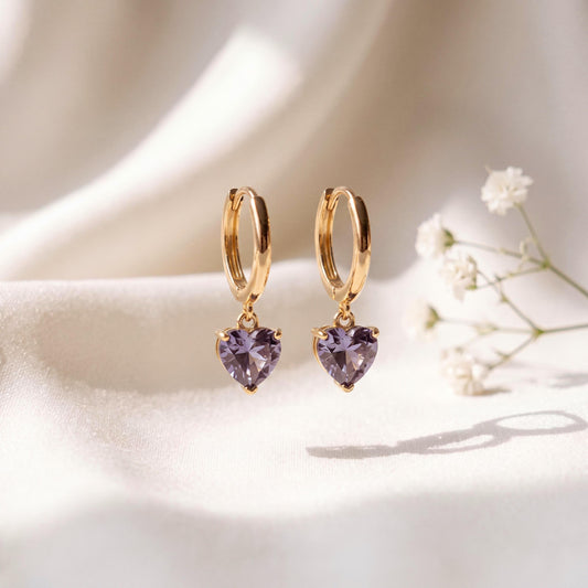 May include: A pair of gold hoop earrings featuring dangling heart-shaped lavender gemstones. The earrings have a polished gold finish and a secure hinged closure, with each heart-shaped stone faceted to catch the light, creating a clear sparkle.