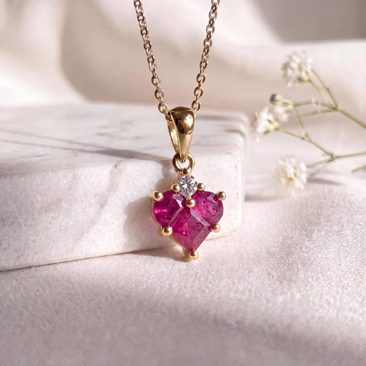 May include: A gold necklace featuring a heart-shaped pendant set with multiple deep pink rubies and a single small clear round gemstone at the top. The pendant hangs from a delicate gold chain, showcasing a polished metal finish and prong settings.