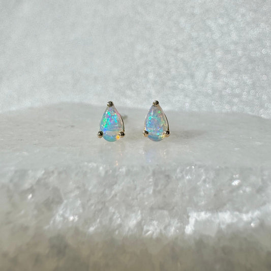 May include: A pair of teardrop-shaped opal stud earrings. The opals display a vibrant play of color, set in gold-colored metal. The earrings are displayed on a white, textured surface.