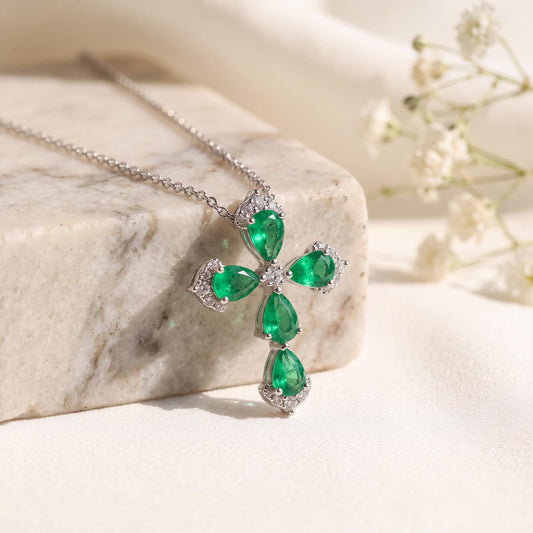 May include: A silver-toned necklace featuring a cross-shaped pendant. The pendant is adorned with four pear-cut green gemstones arranged in a cross pattern, accented by small clear stones along the edges and at the center of the cross.