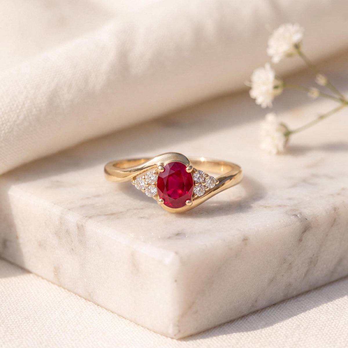 May include: A gold ring featuring a central oval-cut red ruby gemstone flanked by three small round-cut white diamonds on each side. The band has a smooth, polished gold finish that curves gently around the setting of the stones.
