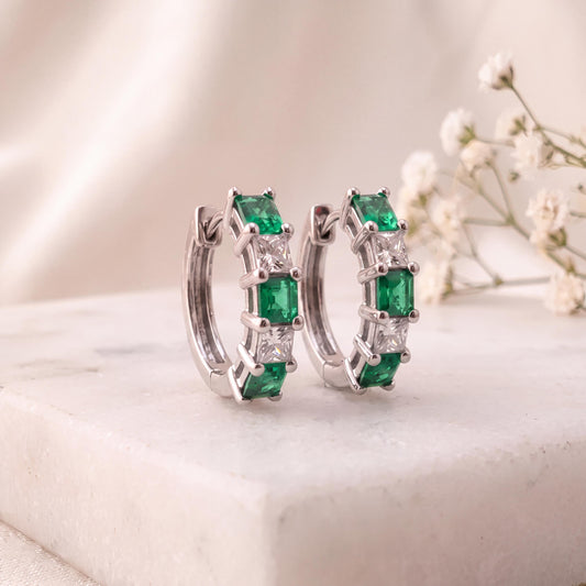 May include: A pair of silver-toned hoop earrings featuring alternating emerald green and clear square-cut stones. The earrings have a hinged closure mechanism and are designed with a row of prong-set gemstones along the front curve of each hoop.
