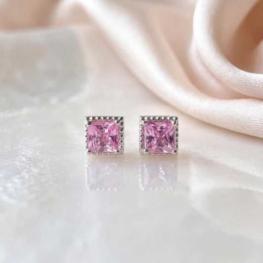 May include: A pair of square-cut pink gemstone stud earrings set in a silver-toned metal frame. Each earring features a faceted pink stone with a brilliant cut, secured by a four-prong setting, reflecting light on a smooth, reflective surface.