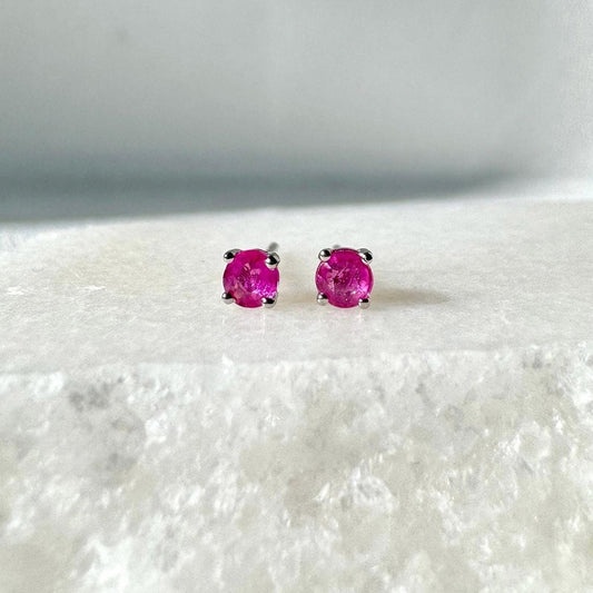 May include: A pair of stud earrings featuring vibrant, round, hot pink gemstones set in silver-colored metal. The earrings are displayed on a textured, white surface, creating a striking contrast.