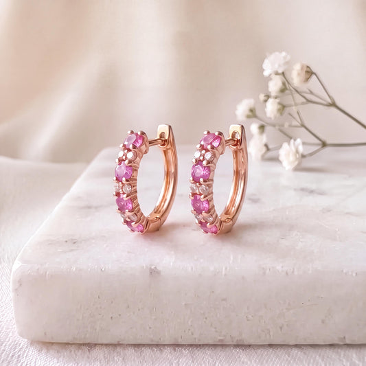 May include: A pair of rose gold hoop earrings featuring a row of alternating round pink and clear stones. The earrings have a hinged closure mechanism and are displayed upright, showcasing the circular design and the vibrant color contrast.