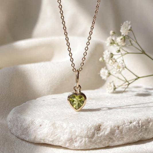 May include: A gold chain necklace featuring a small heart-shaped pendant with a faceted light green peridot gemstone set in a smooth gold frame. The pendant hangs from a delicate gold link chain, showcasing a clear, bright green stone.